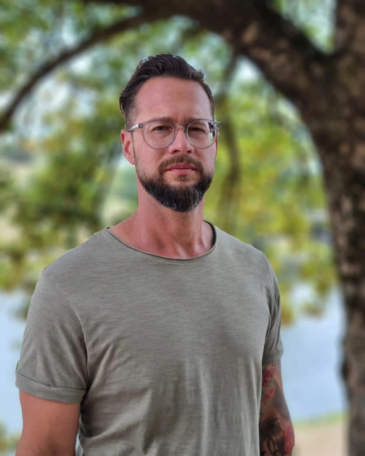 Der Inhaber im Porträt, draußen unter einem Baum, olivgrünes T-Shirt, Brille und Bart, Blick in die Kamera